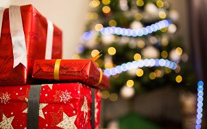 trzy prezenty świąteczne opakowane w czerwony papier ozdobny, przewiązane różnokolorowymi wstążkami, z tyłu niewyraźnie widoczna drzewko choinkowe z jasnym łańcuchem świetlnym i żółtymi światełkami